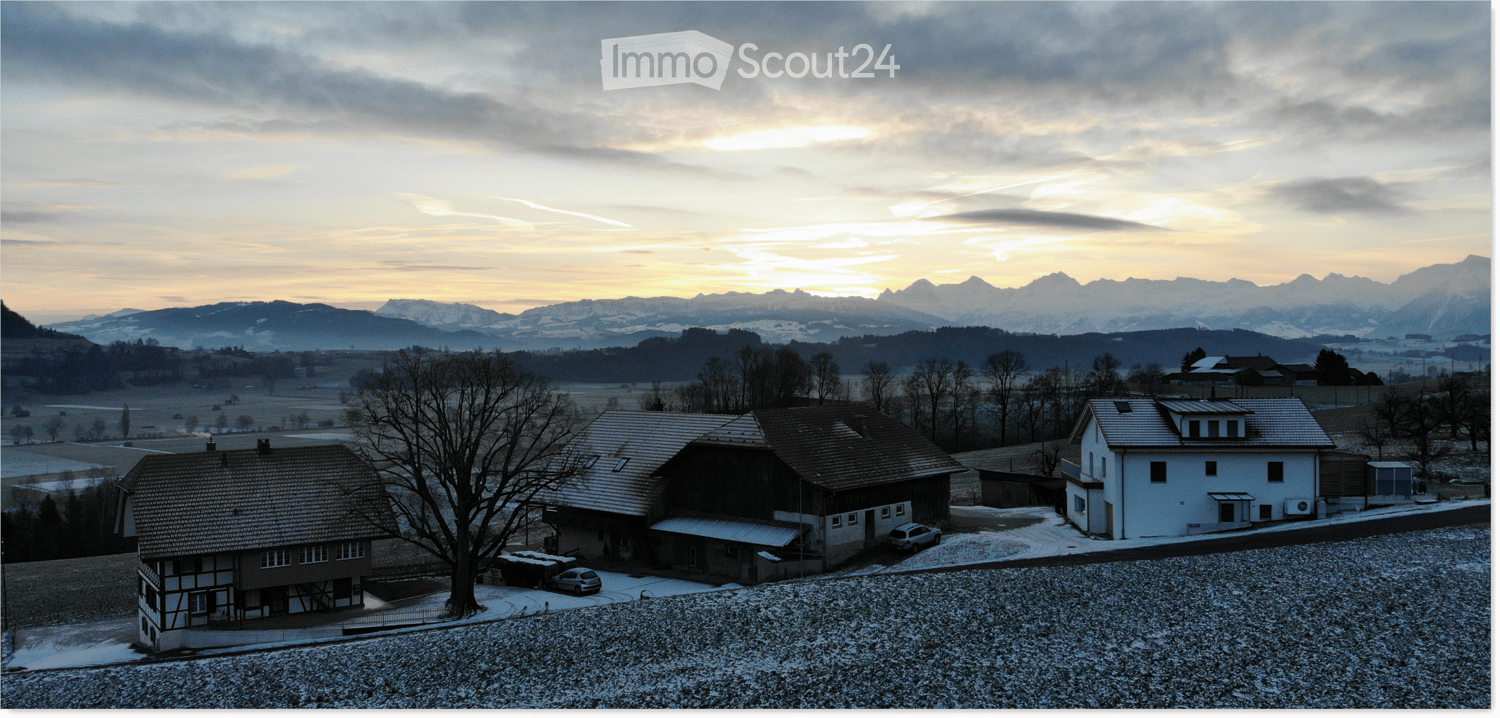 Aerial view of a house with a tiled roof, surrounded by a snow-covered yard and trees, with a scenic mountain backdrop at sunset.