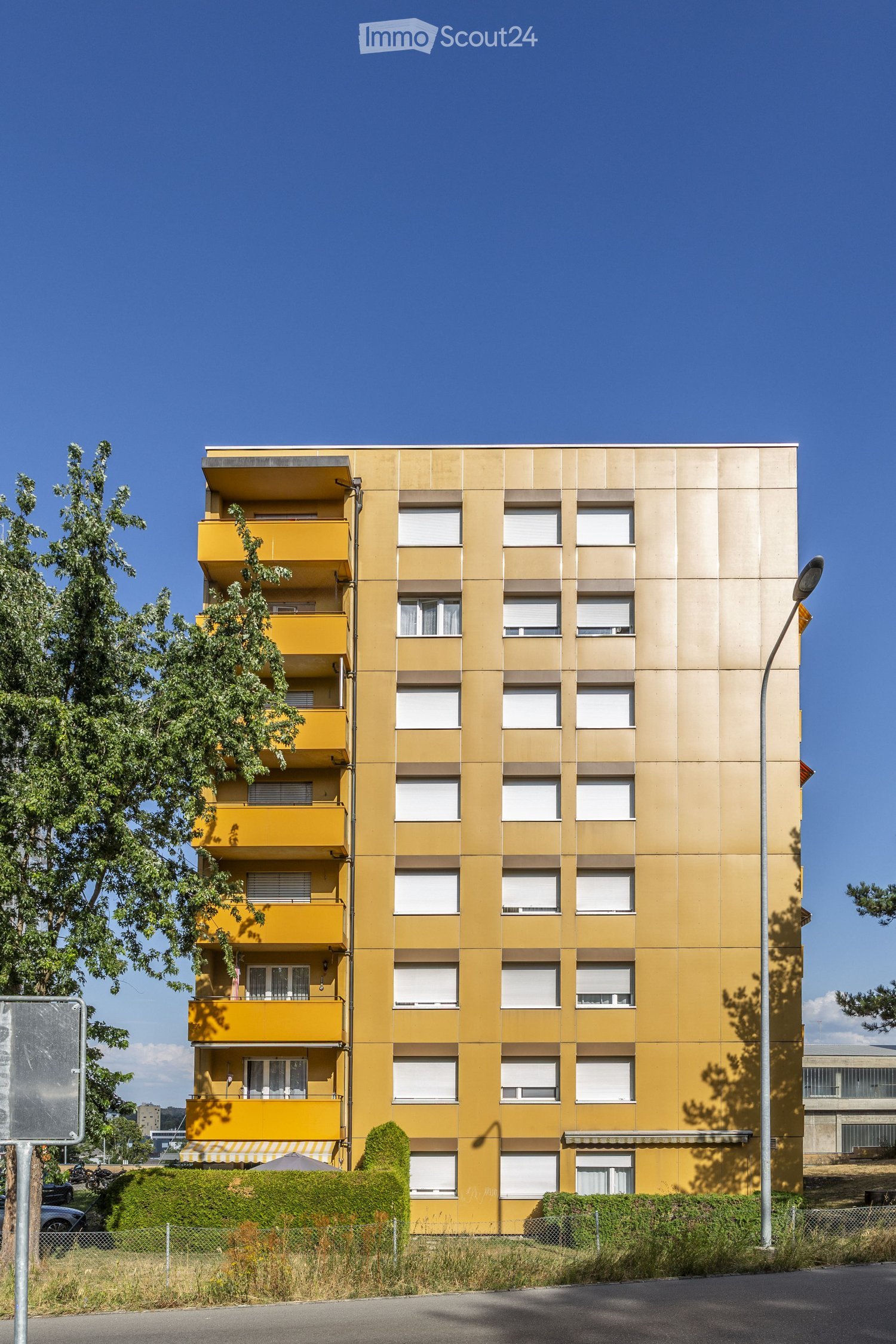 A multi-story residential building with yellow balconies and white-framed windows, surrounded by greenery and a clear blue sky.