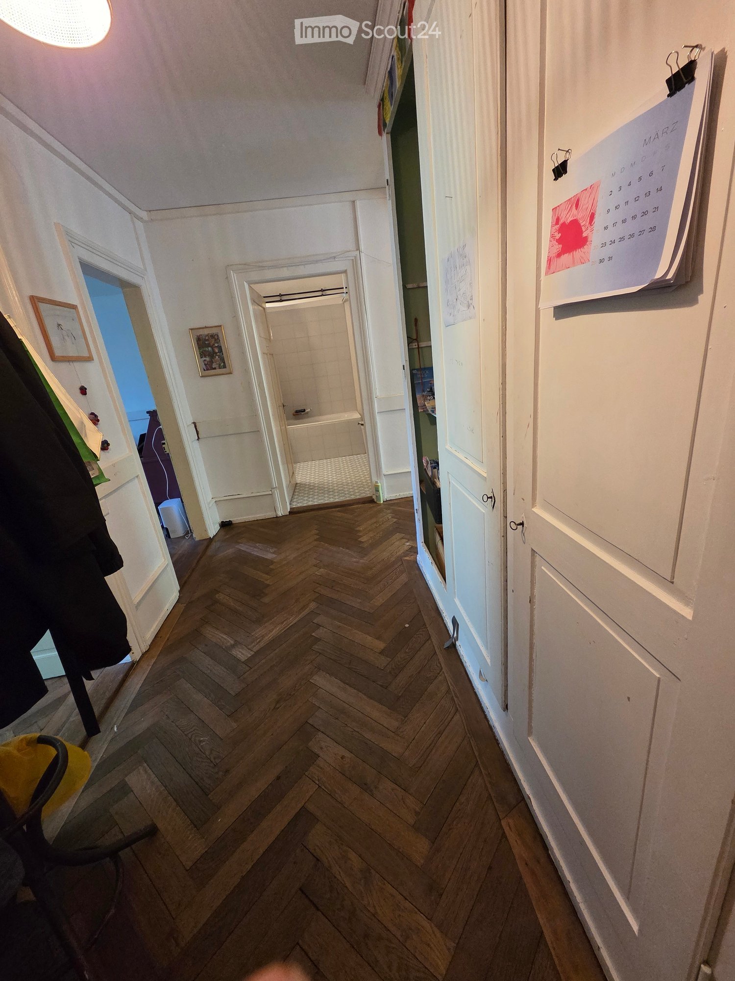 Hallway with herringbone wooden flooring, white walls, and doors leading to rooms. Calendar and coat rack visible.