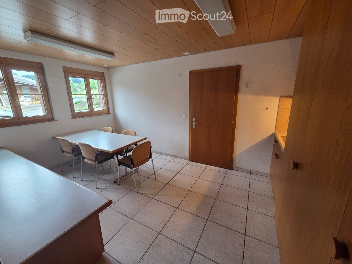 Bright room with tiled floor, white walls, and wooden ceiling. A white table with four chairs is positioned near the windows, and a wooden door leads to another area.