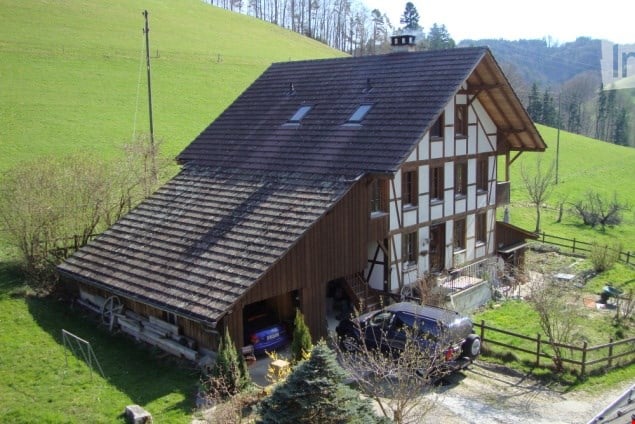A two-story house with a wooden exterior and dark roof, situated on a hillside with a garage and parking area.
