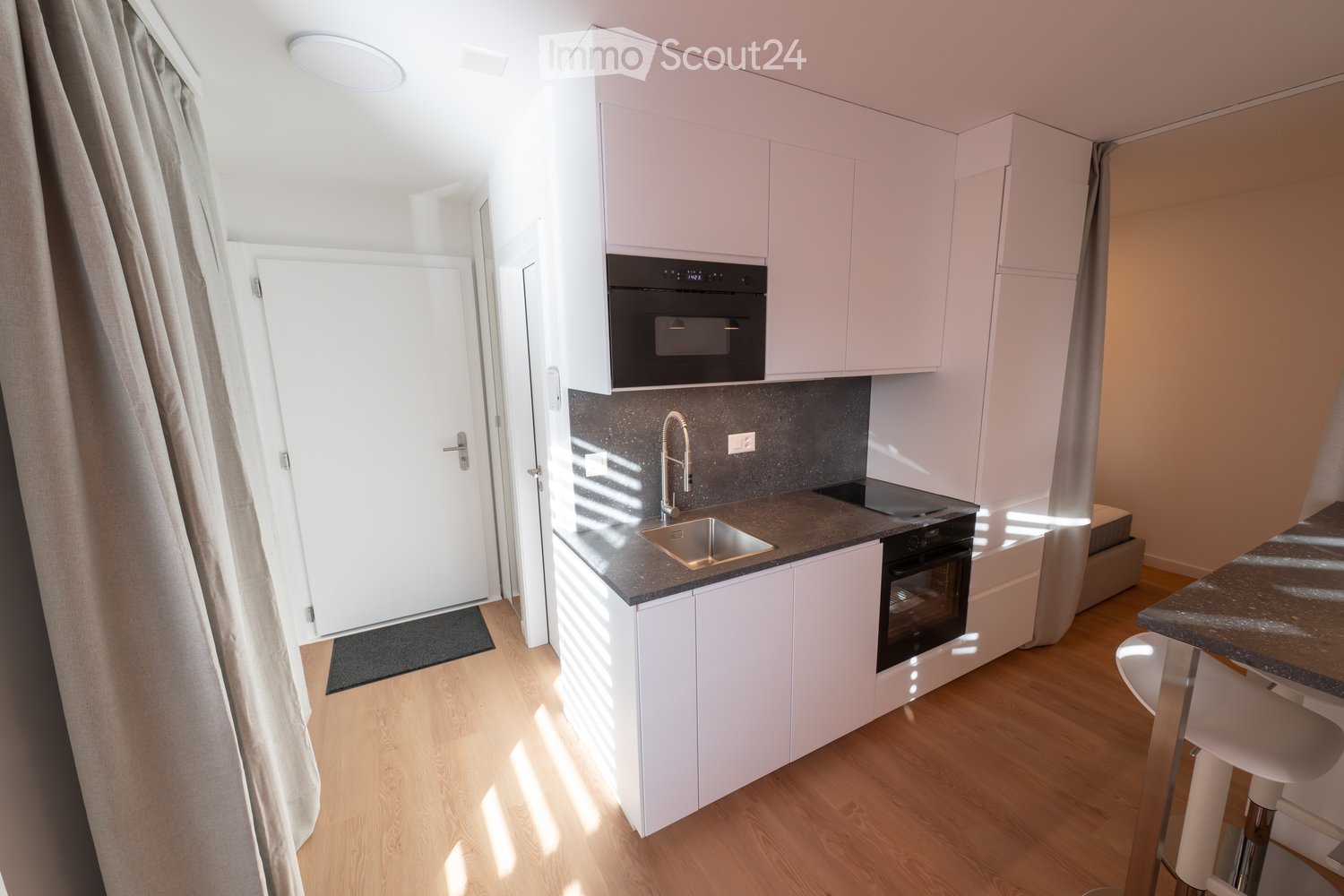 Modern kitchen with white cabinets, dark countertops, stainless steel sink, microwave, and oven. Wooden flooring and a small seating area with a stool.