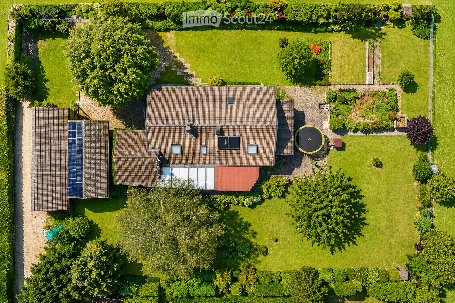 Aerial view of a two-story house with a tiled roof, surrounded by a well-maintained garden with a trampoline, solar panels on the roof, and a paved driveway.
