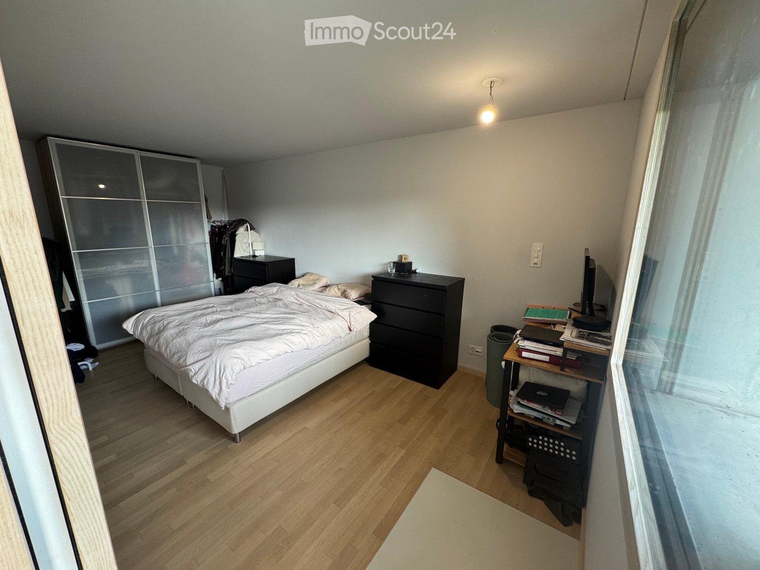 A modern bedroom with a wooden floor, a white bed with beige bedding, a black nightstand, a wardrobe, and a small table with books and a laptop.