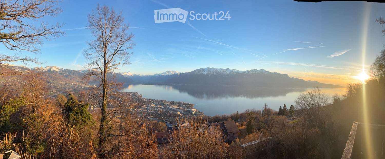 Panoramic view of a lake and snow-capped mountains, with a town in the foreground and trees on the hillside.