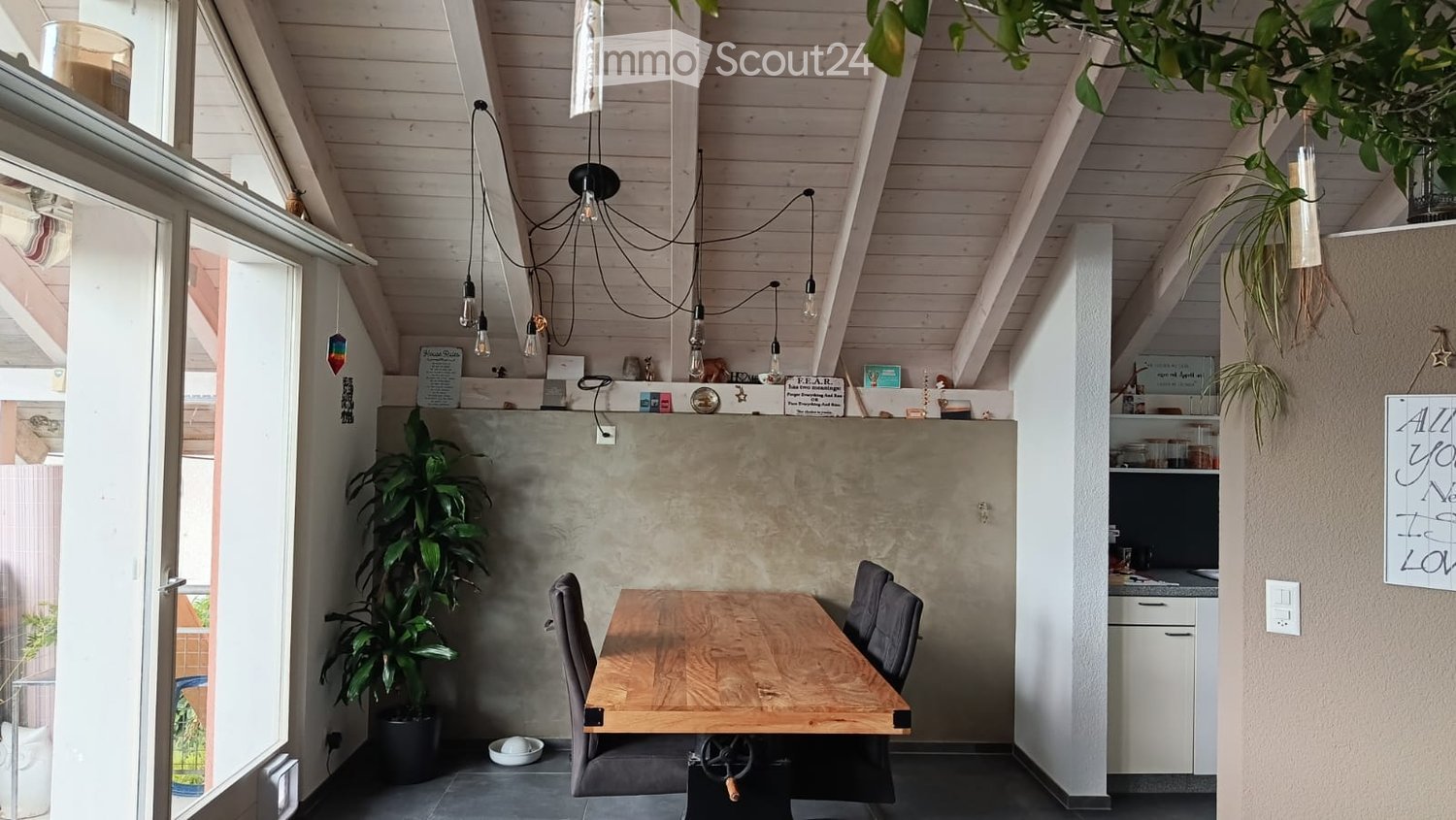 Modern, open-plan room with a wooden dining table, chairs, kitchen area, pendant lights, and plants.