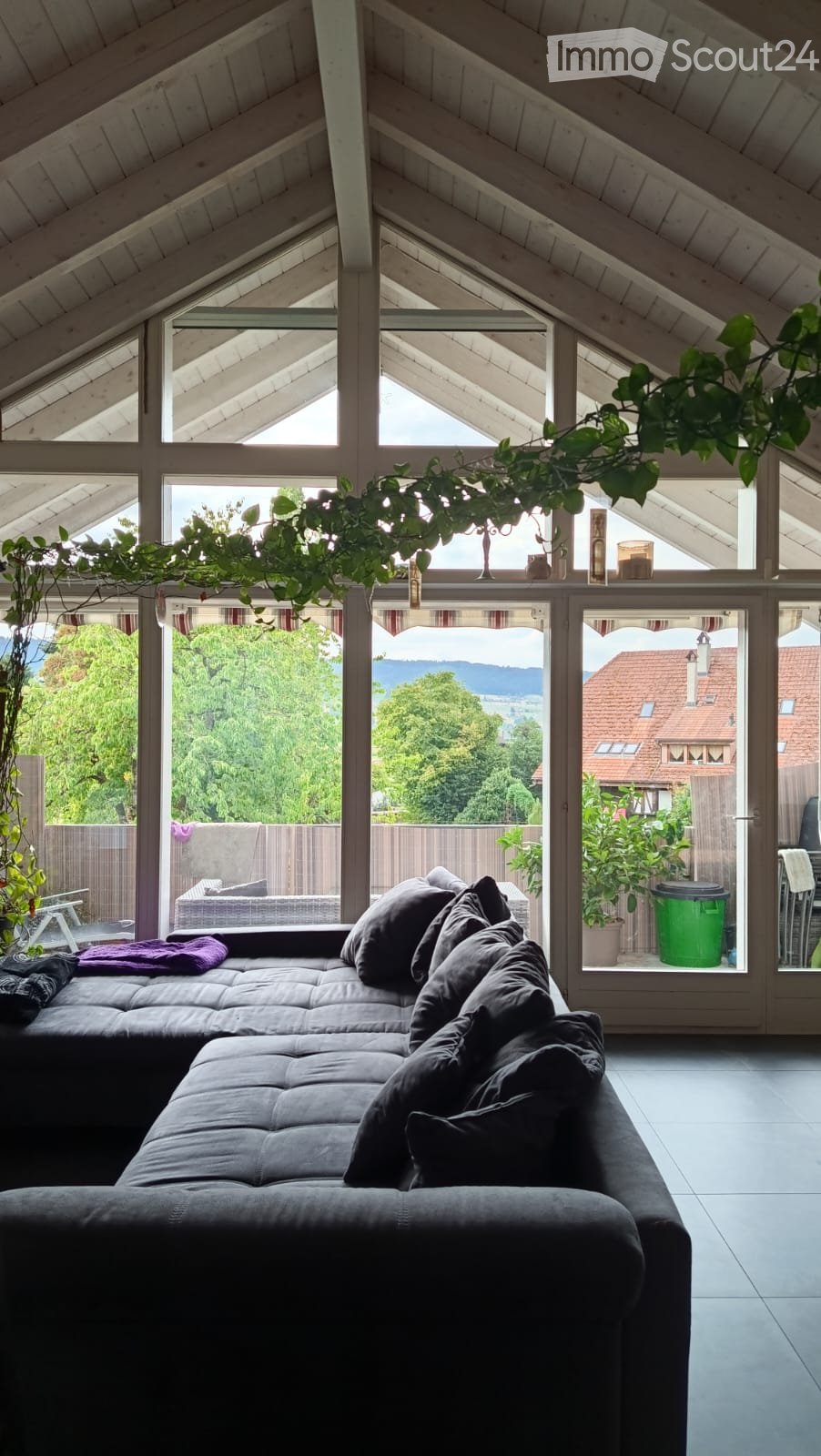 Bright living room with a grey sectional sofa, multiple pillows, a small table, and a hanging plant. Large windows offer a scenic view of the outdoors.