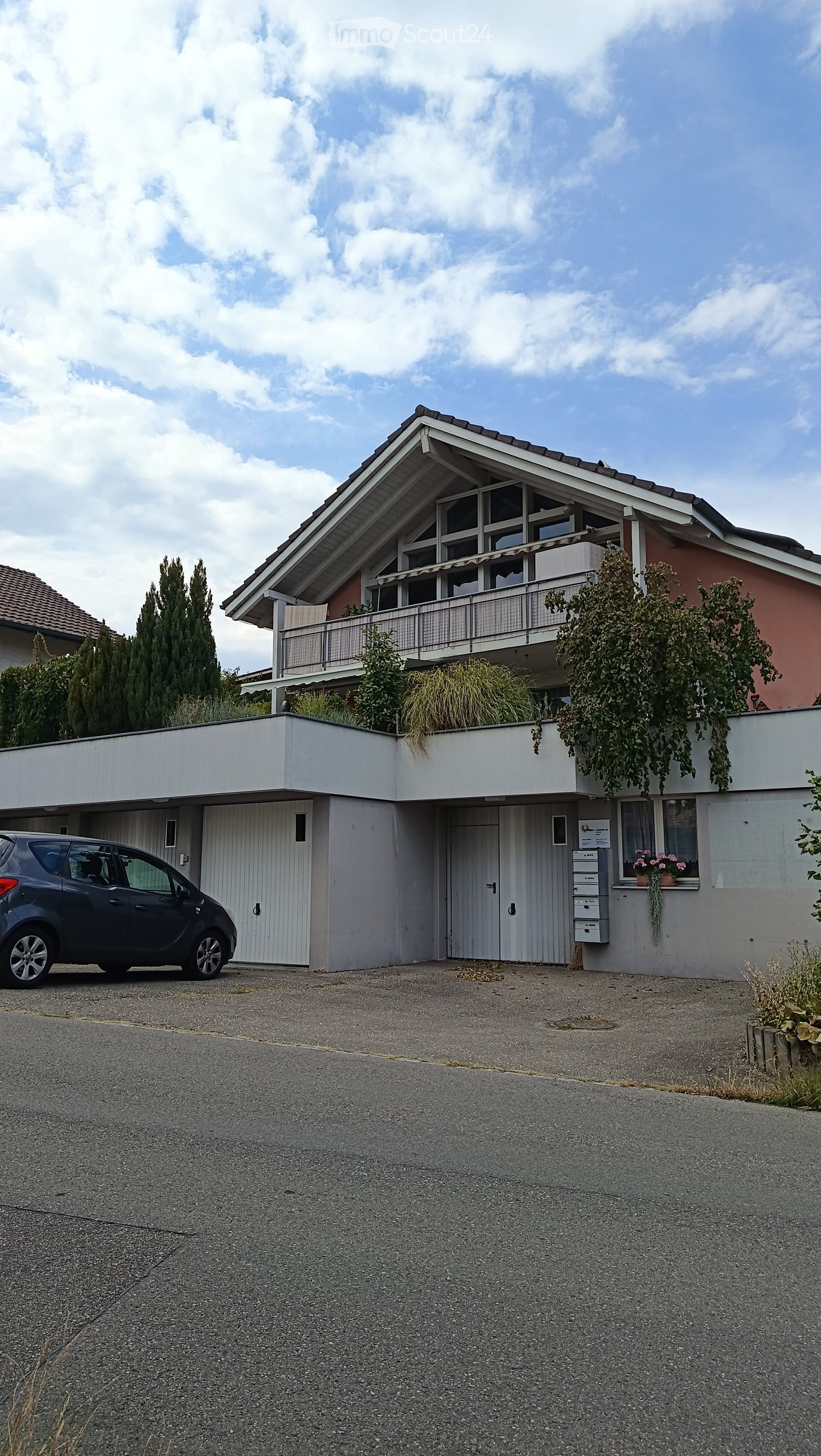 Two-story house with a modern design, featuring a flat roof, large windows, and a balcony on the upper level. The house has a garage and is surrounded by greenery.