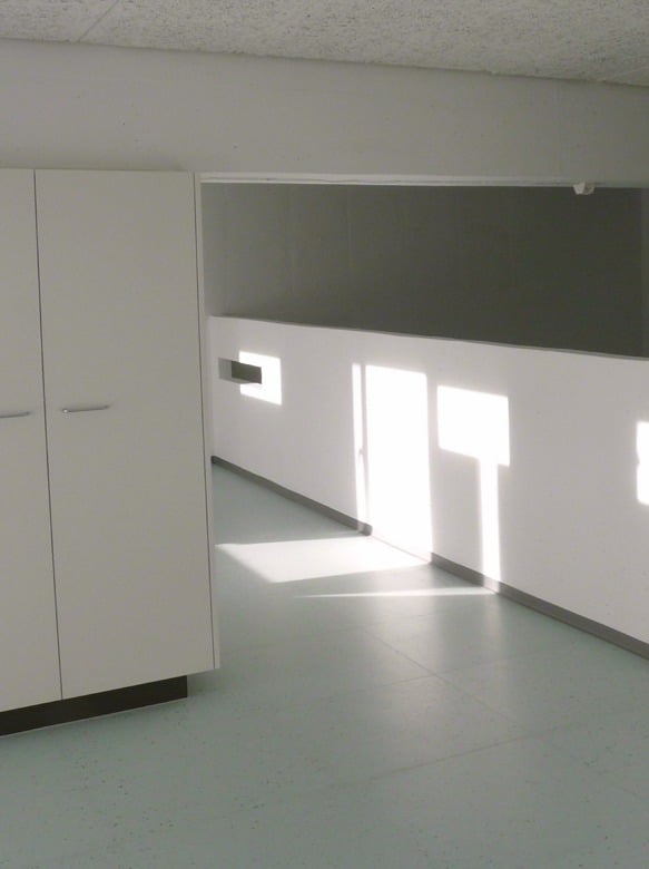 Empty kitchen area with white cabinets and a tiled floor, illuminated by natural light from a window.