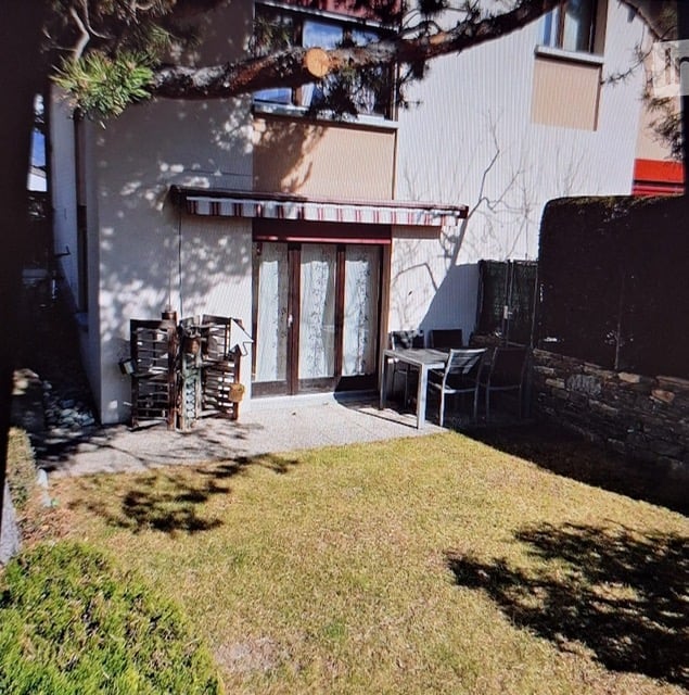Small garden area with a patio, outdoor furniture, and a wooden shed. The house has a modern facade with large windows and a covered entrance.