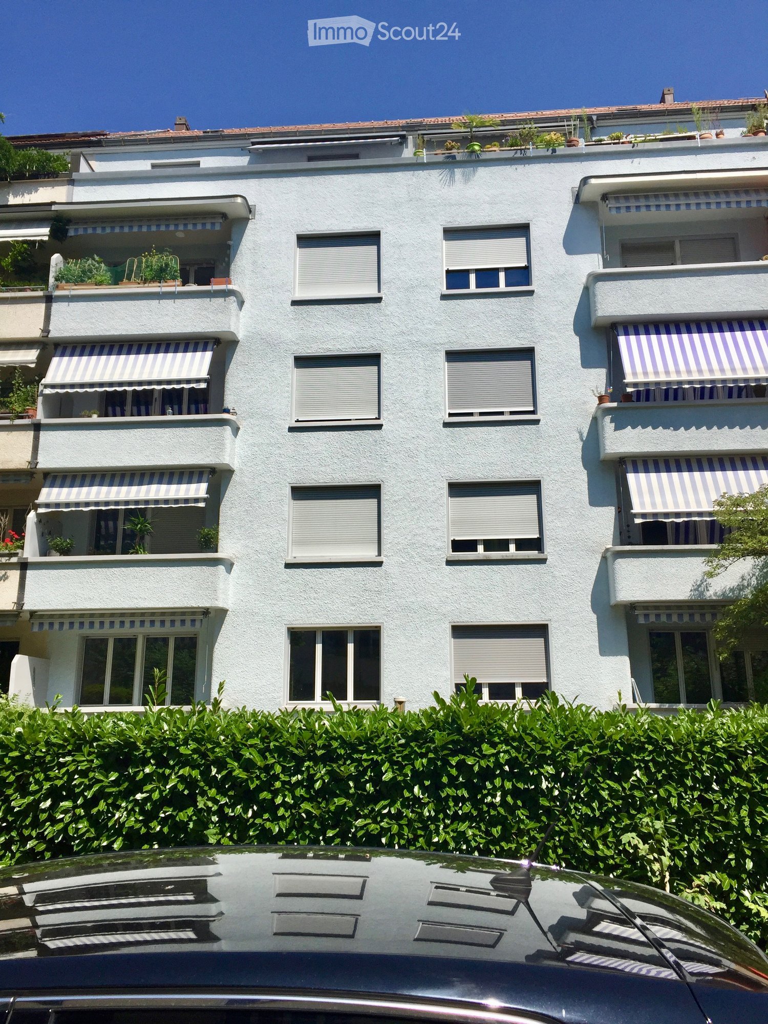 Four-story residential building with white facade, multiple windows, balconies with striped awnings, and rooftop greenery.