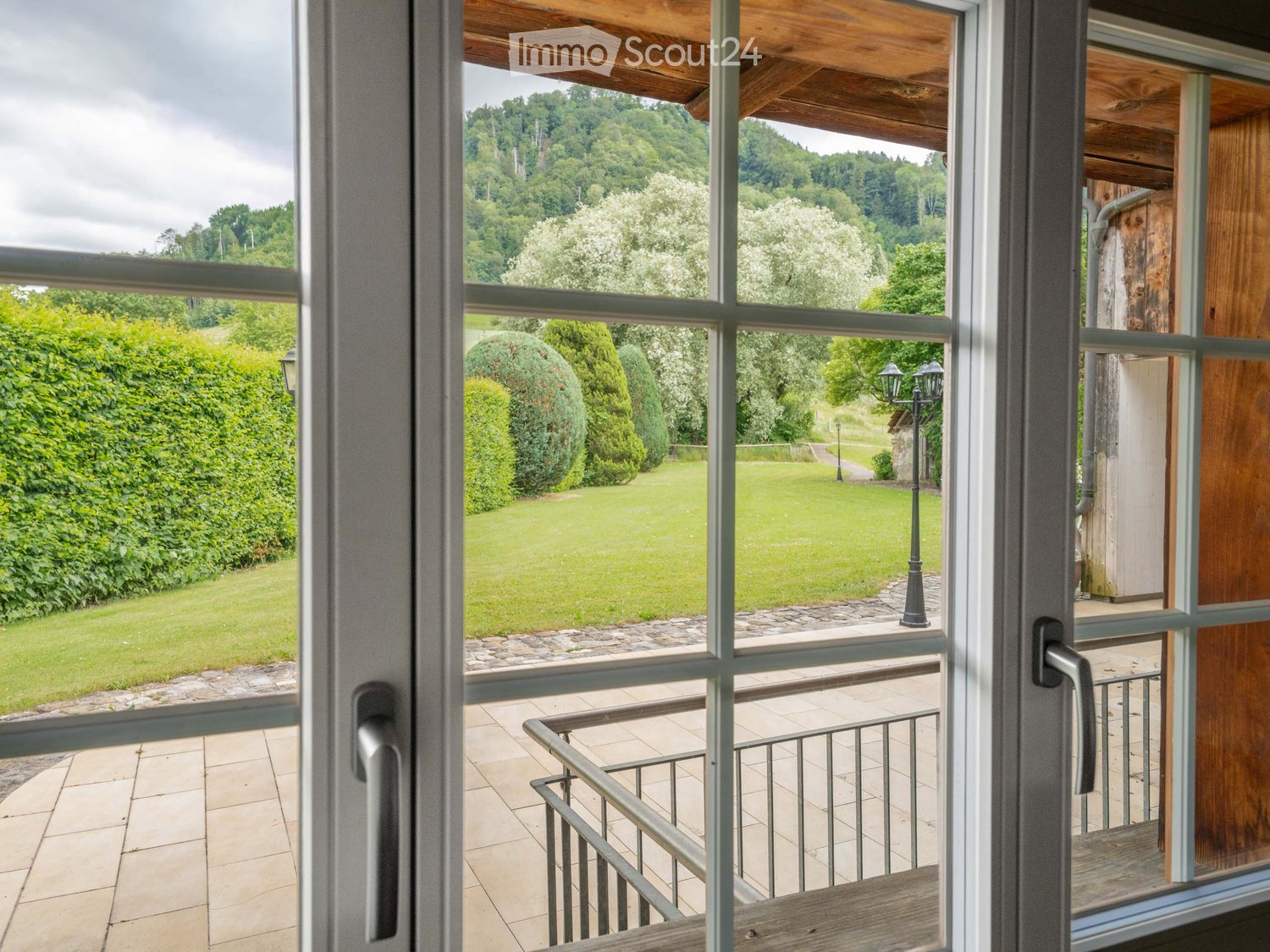 Balcony with wooden decking, metal railing, and scenic view of a green lawn, trees, and hills.