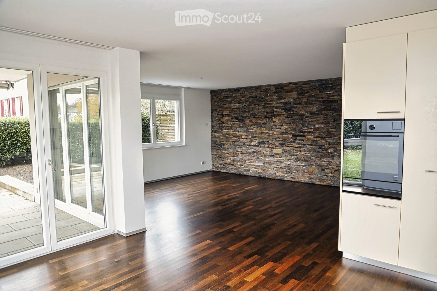 Modern kitchen with white cabinets, a stone accent wall, and a dark wooden floor. Features a built-in oven and large sliding glass doors leading to an outdoor area.