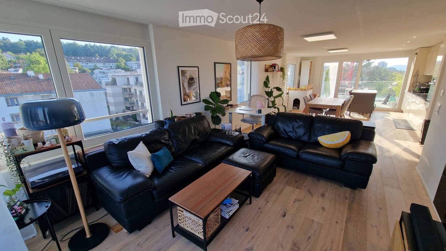 Modern living room with large windows, black leather sofa, coffee table, floor lamp, and dining area visible in the background.