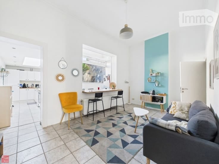Bright, modern living space with a grey sofa, coffee table, yellow accent chair, and small kitchen area with bar stools. Features a geometric patterned rug and white tiled flooring.