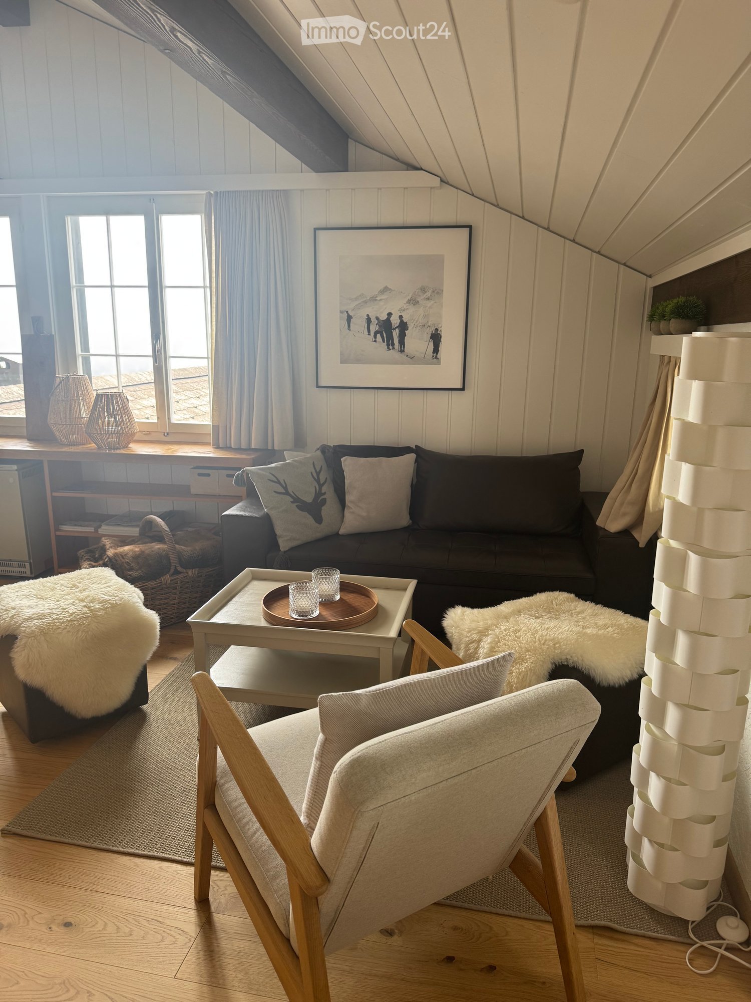 Cozy attic living room with slanted wooden ceilings, large windows, a dark grey sofa with decorative pillows, a wooden coffee table with glass drinks, a light grey armchair, and a fluffy white throw on a side table.