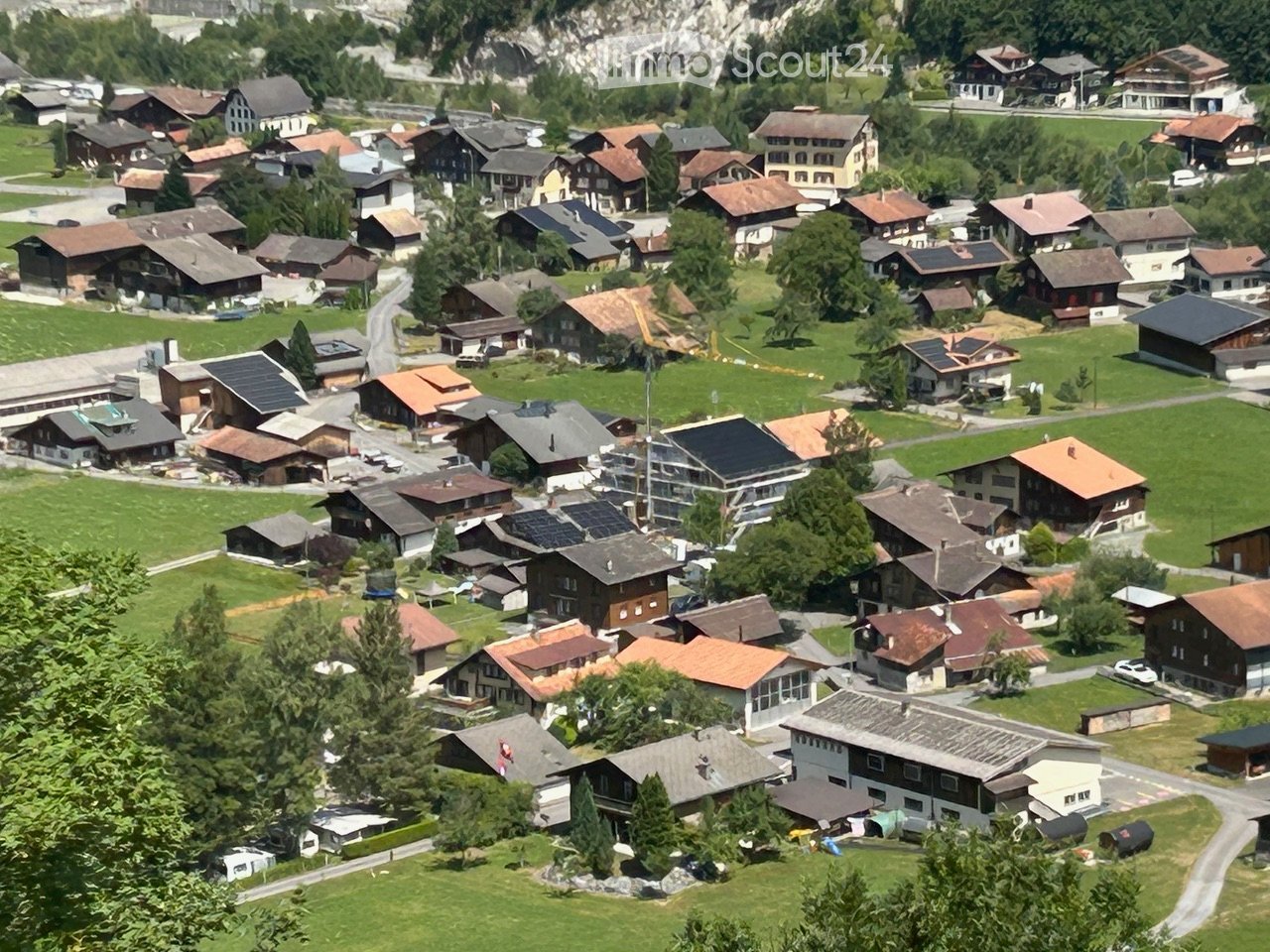 Aerial view of a small village with numerous houses, green lawns, and scattered trees. The houses have varied roof styles and colors, and the area is surrounded by mountains.