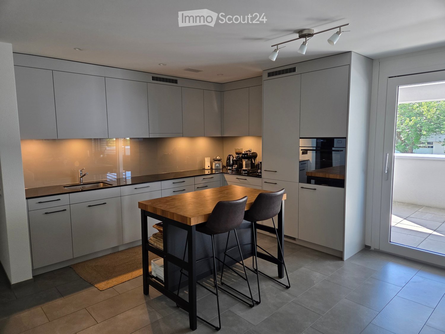 Modern kitchen with grey cabinets, a wooden island table, two stools, a sink, and a coffee maker on the counter.