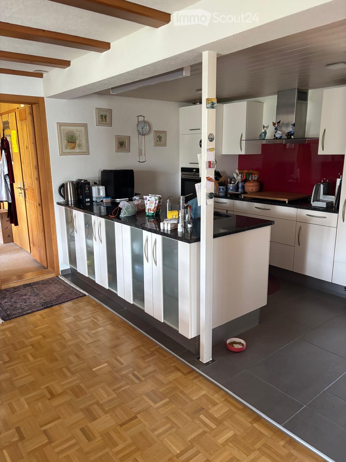 Modern kitchen with white cabinets, black countertops, wooden herringbone flooring, and various appliances including a coffee maker, microwave, and oven. A pet bowl is visible on the floor.