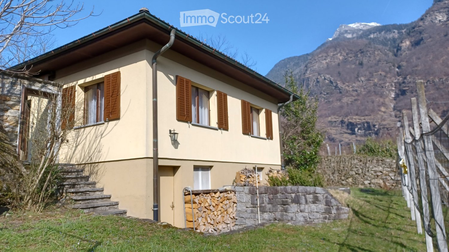 Two-story house with beige walls, brown shutters, stone steps, and a stack of firewood near the entrance, set against a mountainous backdrop.