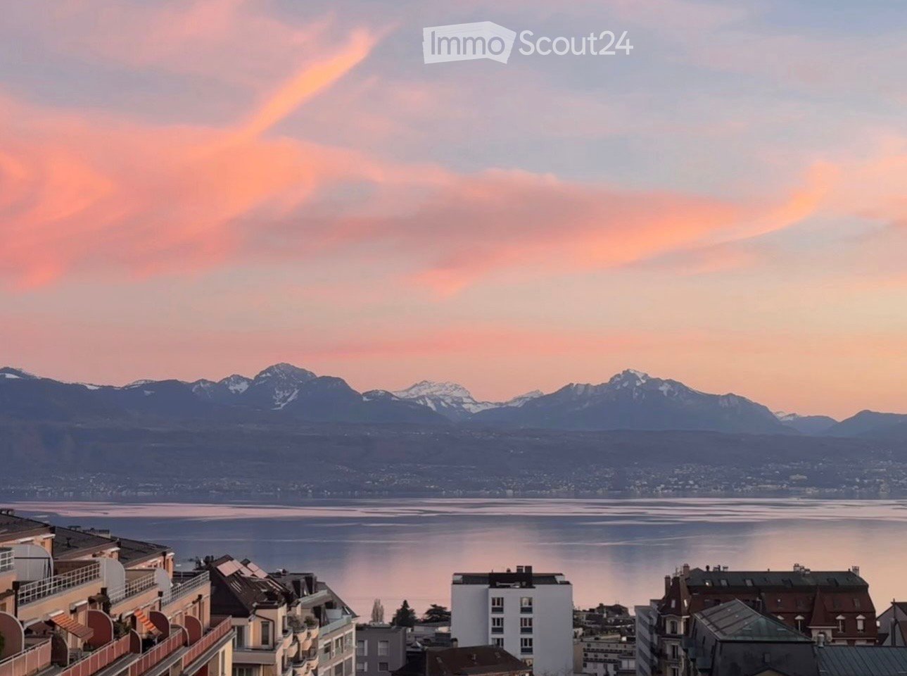 Panoramic view from a balcony or terrace overlooking a lake and distant mountains during sunset.