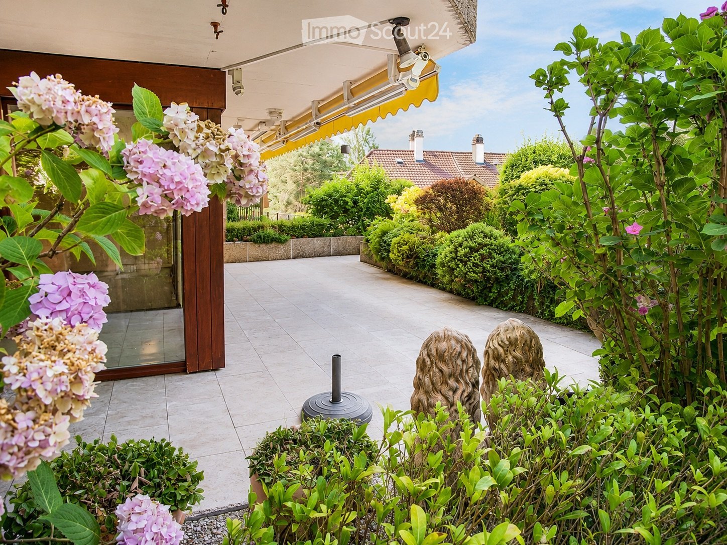 Well-maintained garden with lush greenery, flowering shrubs, decorative statues, and a covered patio area with a glass door leading inside.