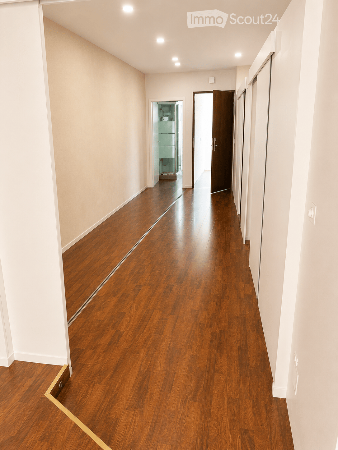 Empty hallway with polished wooden flooring, white walls, and recessed lighting. Features a partially open door leading to a bathroom and another closed door on the right side.