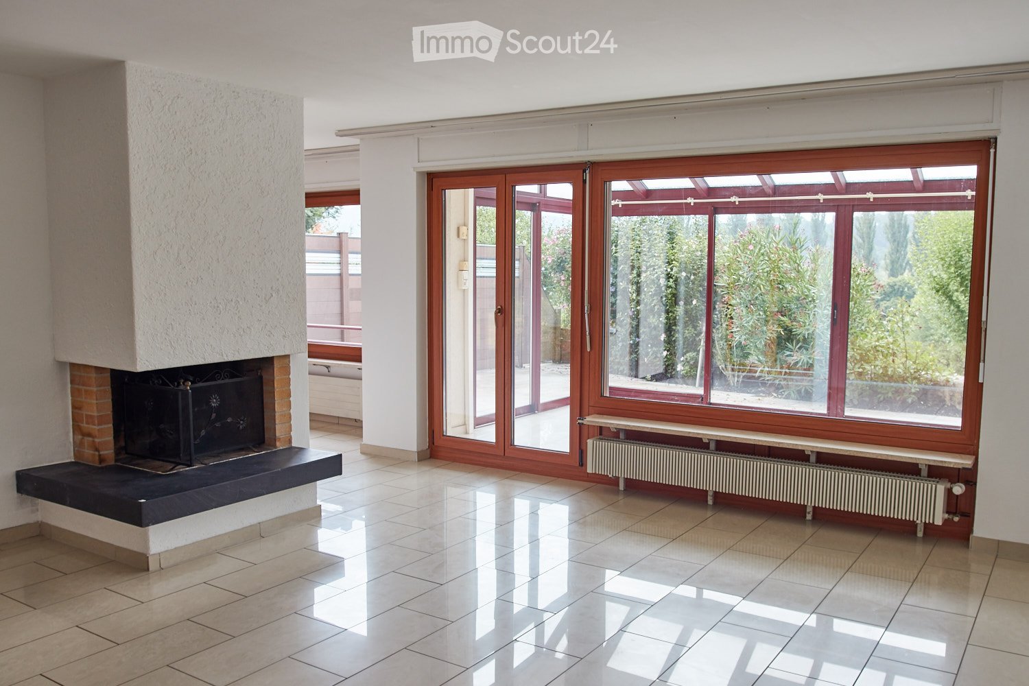 Empty living room with tiled floor, large windows, fireplace, radiator, and sliding glass doors leading to a balcony.