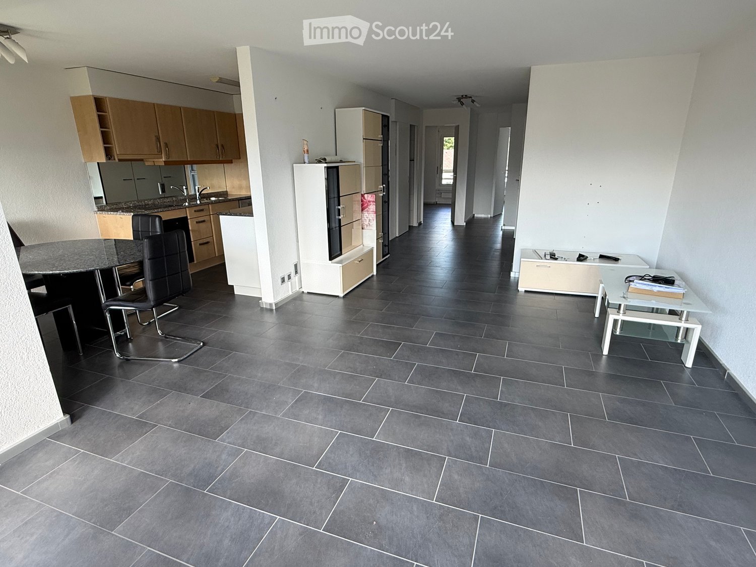 Open-plan living and dining area with grey tiled flooring, modern kitchen area with cabinets and appliances, and a dining table with chairs.