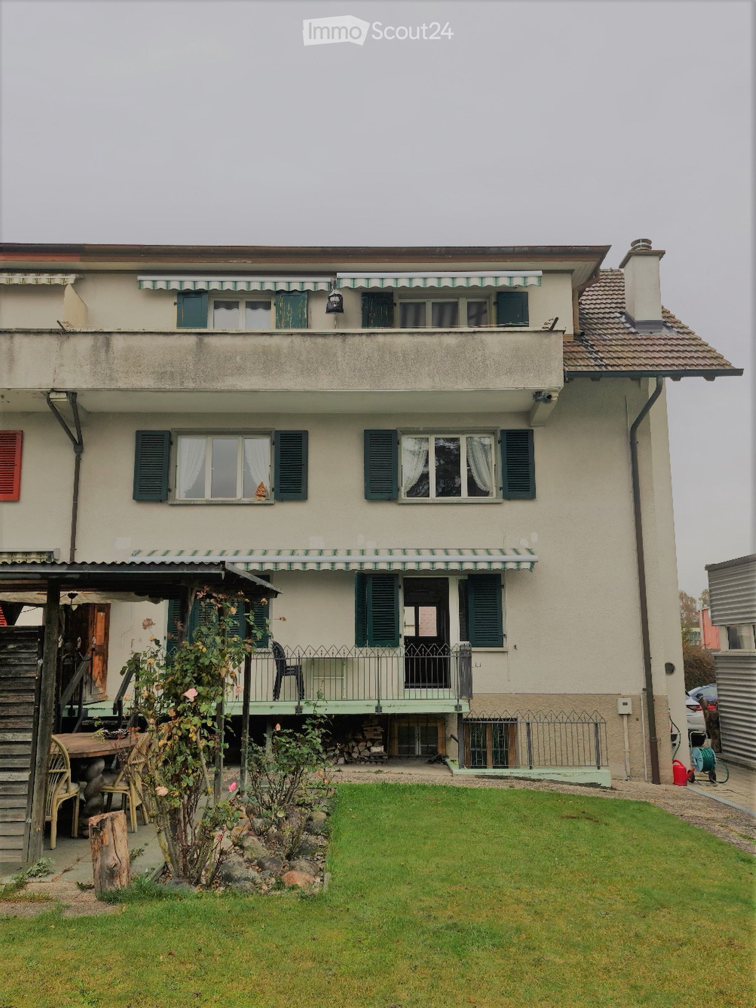 Two-story house with beige walls, green shutters, and a balcony with striped awnings. The front yard features a well-maintained lawn, a wooden staircase leading to a patio area with outdoor furniture, and a small garden with plants and a wooden table.