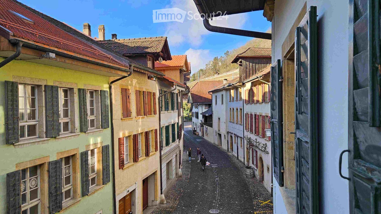 A narrow cobblestone street lined with colorful, multi-story buildings with traditional European architecture, featuring wooden shutters and tiled roofs.