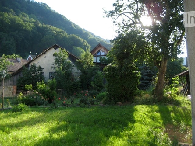 A peaceful garden area with a well-maintained lawn, surrounded by trees and shrubs. Two houses are visible in the background, nestled among greenery and hills.