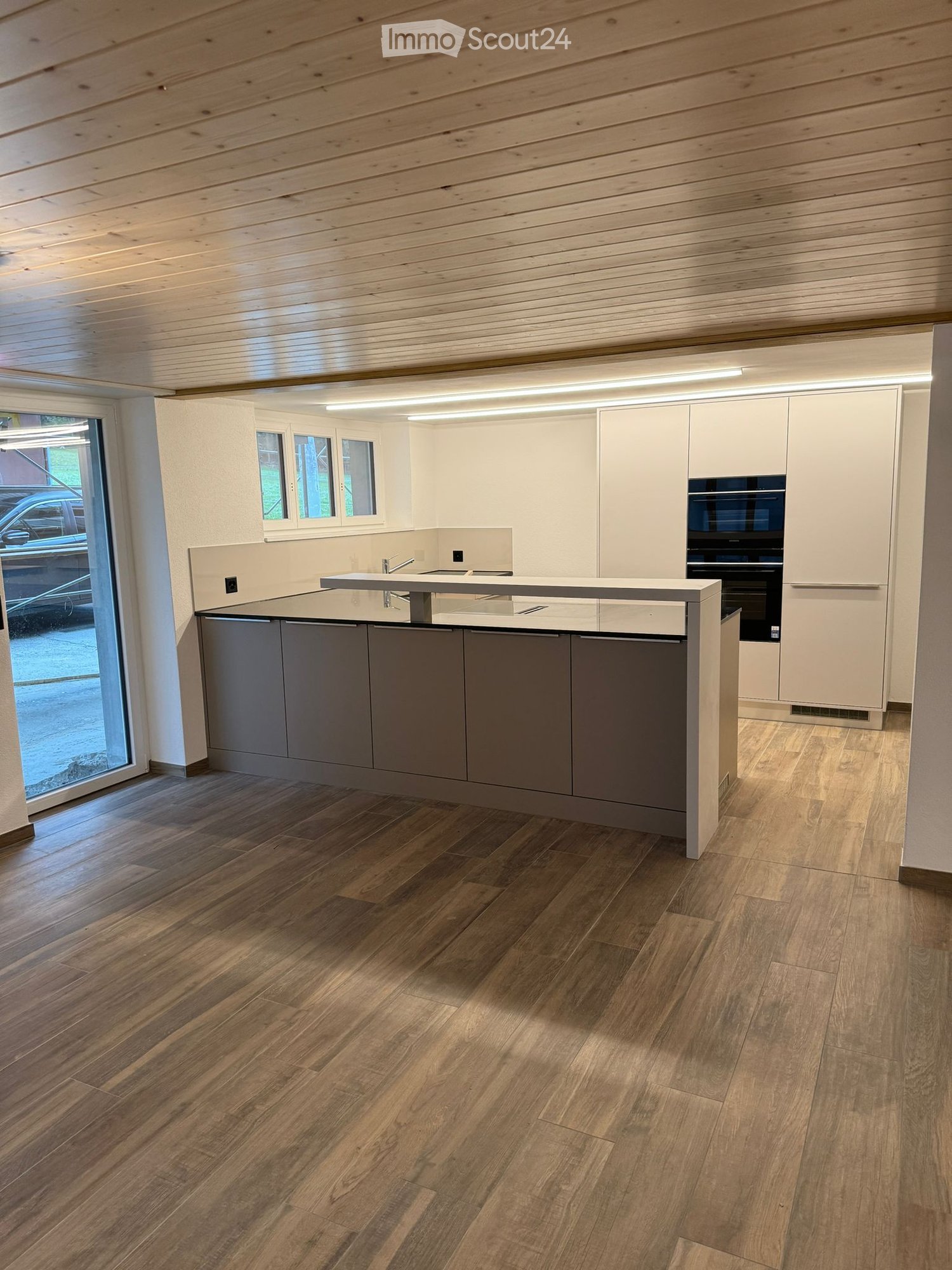 Modern kitchen with light wood ceiling, wooden flooring, grey cabinets, white upper cabinets, and a central island with a sink and faucet.