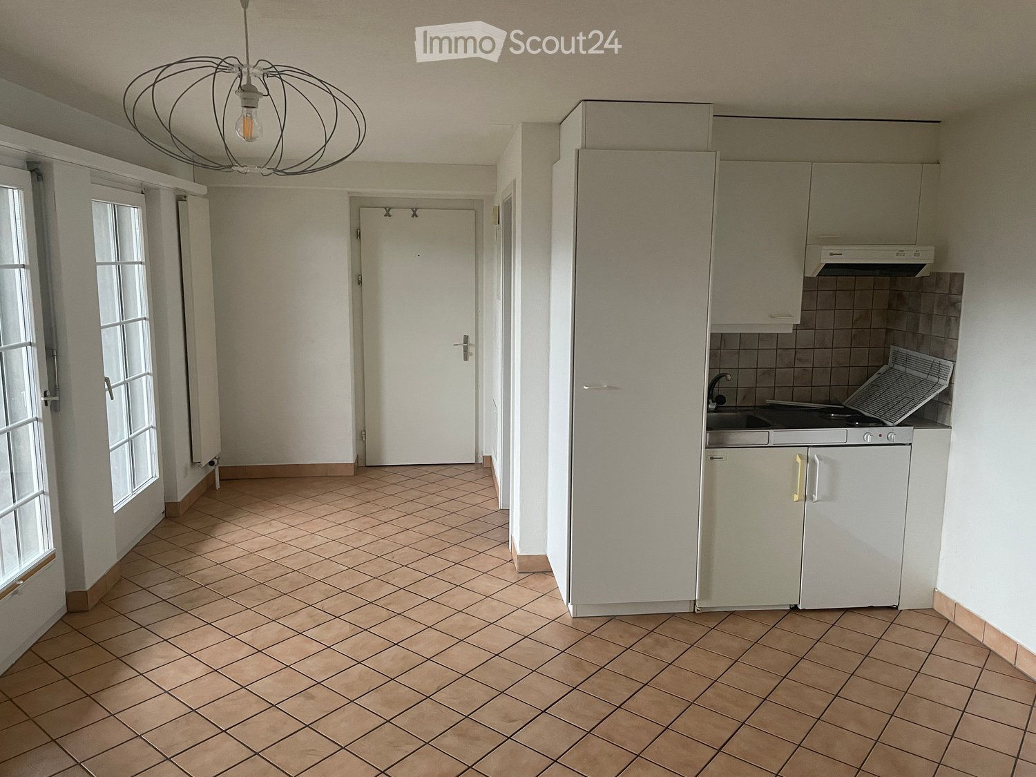 Empty room with tiled floor, white walls, and a small kitchen area with cabinets and a sink.