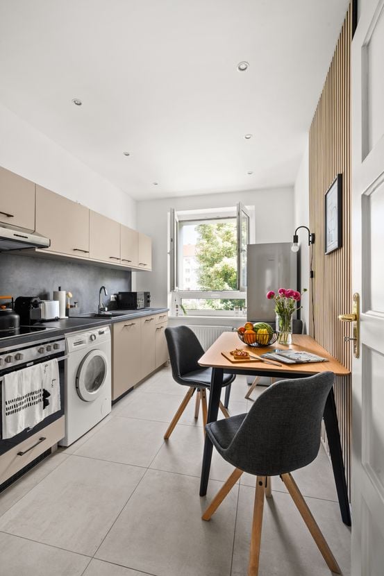 Modern kitchen with light-colored cabinets, stainless steel appliances, a central island, and a small dining area with a table and chairs. The space is bright with large windows and includes a fruit bowl and vase on the table.
