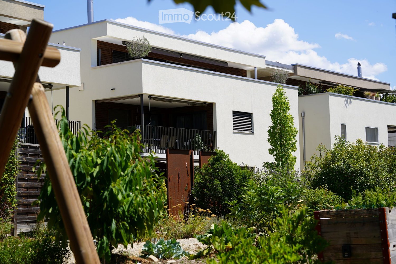 Modern two-story building with white facade, large windows, and a balcony. The surrounding area includes a garden with various plants and a wooden structure, likely part of a residential complex.