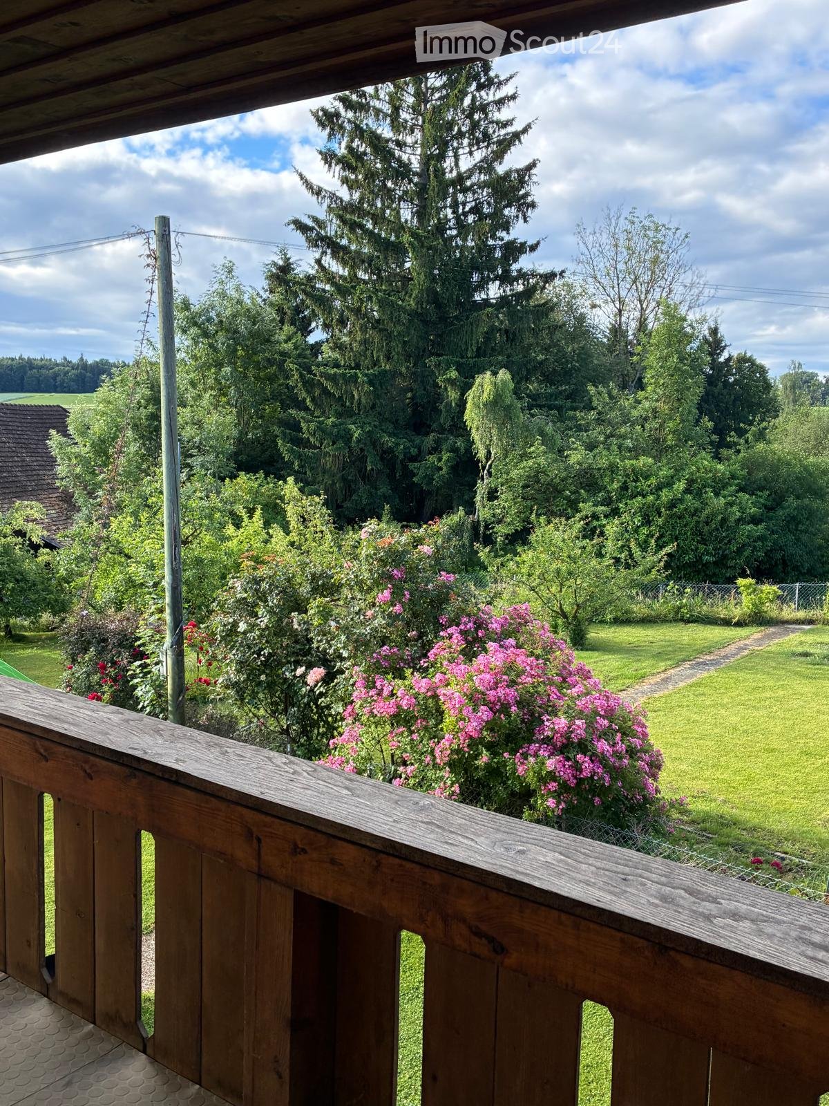 Wooden balcony overlooking a lush garden with pink flowers, green grass, and tall trees under a partly cloudy sky.