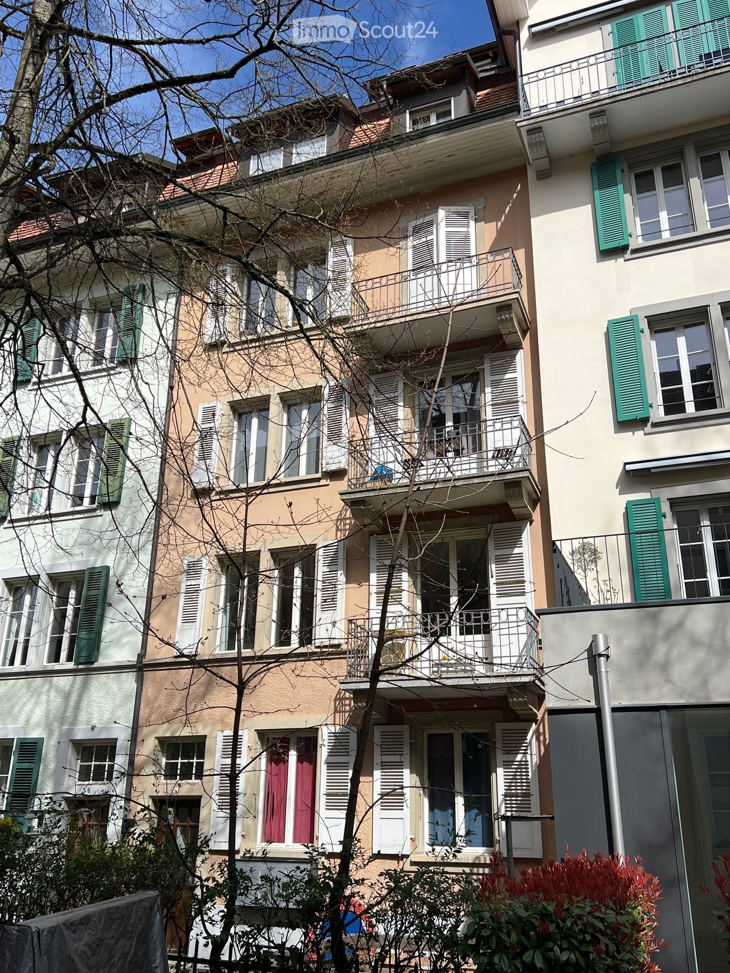 Multi-story residential building with balconies, green shutters, and a tree in front.