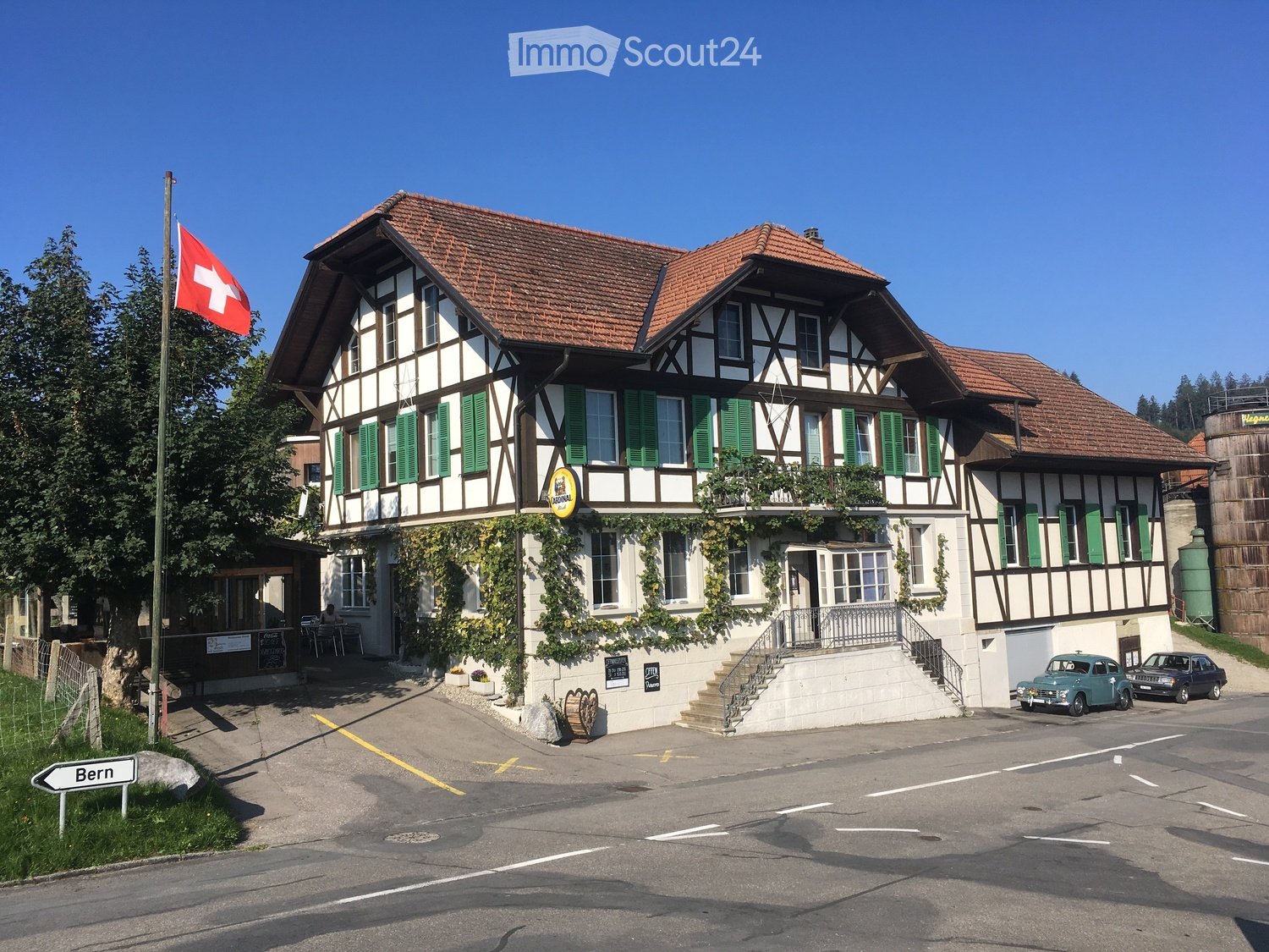 Traditional Swiss building with a red-tiled roof, green shutters, and a balcony adorned with plants. A Swiss flag is displayed on a pole, and a sign reading "Bern" is visible on the left side.