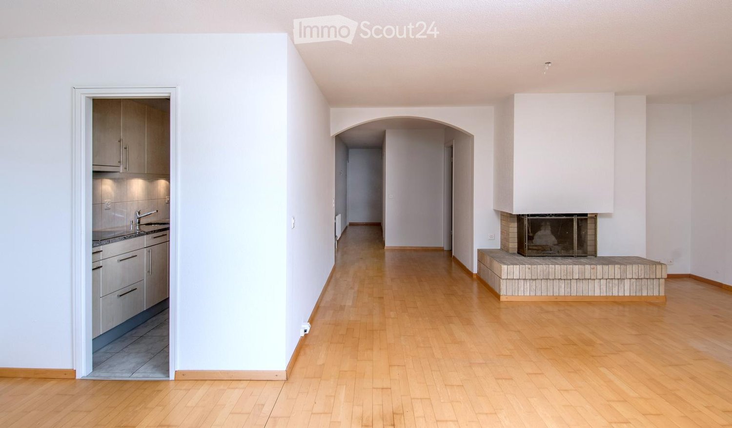 Empty living space with light wooden flooring, white walls, and a fireplace with a tiled surround. A small kitchen area is visible through an open doorway.