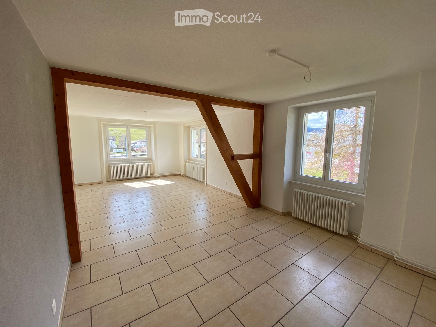 Empty room with tiled floor, white walls, and large windows. Radiators are visible along the walls.