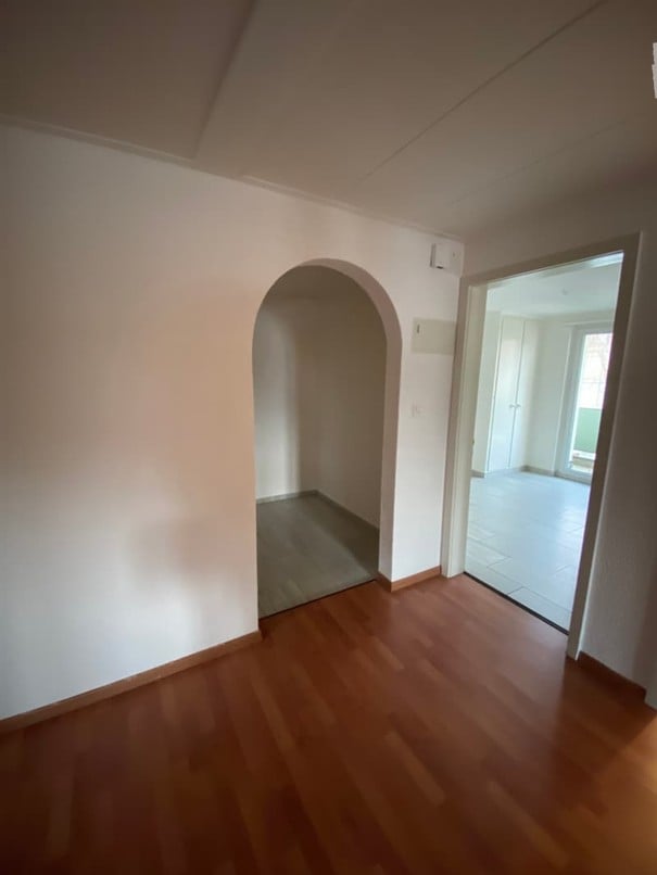 Empty room with wooden flooring and white walls, featuring an arched doorway leading to another room.
