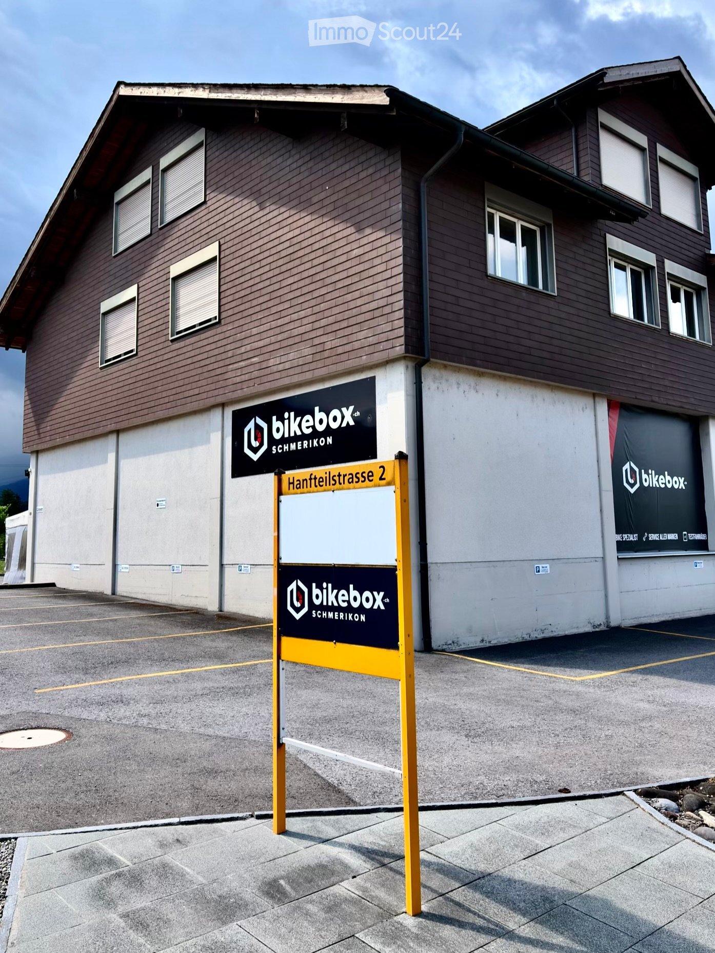 Two-story building with brown wooden facade, multiple windows, and signage for "bikebox.ch" and "SCHMERIKON" on the walls. A yellow signpost in front displays the address "Hantteilstrasse 2".