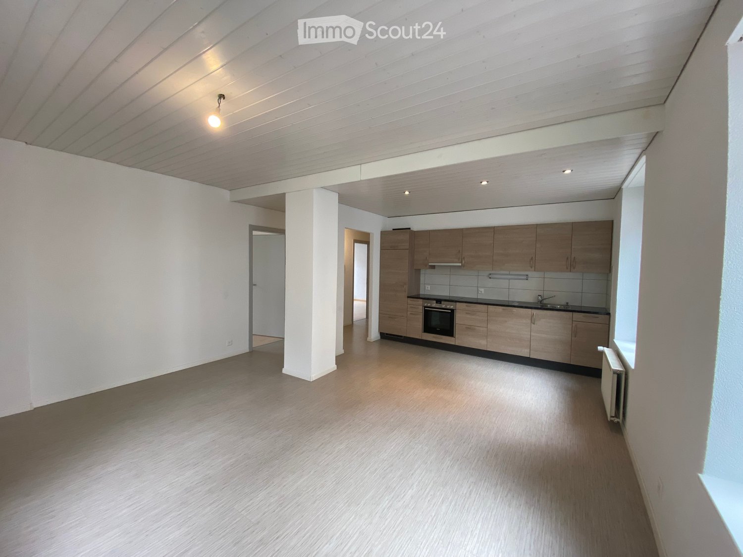Bright, unfurnished living space with wooden flooring, white walls, and a modern kitchen area featuring wooden cabinets, an oven, and a sink.