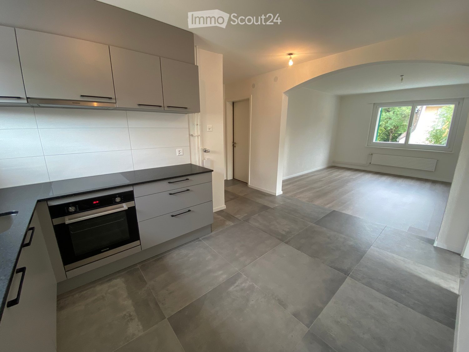 Modern kitchen with grey cabinets, stainless steel oven, and tiled floor. Adjacent unfurnished room with wooden flooring and large window.