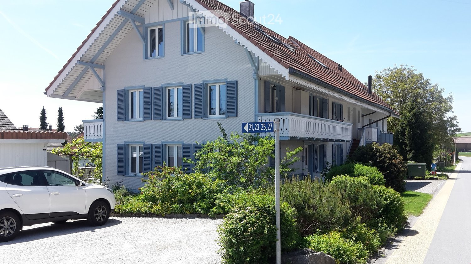 Two-story house with white walls, blue shutters, and a red-tiled roof. Features a balcony on the upper floor and is surrounded by greenery and a driveway with a parked car.