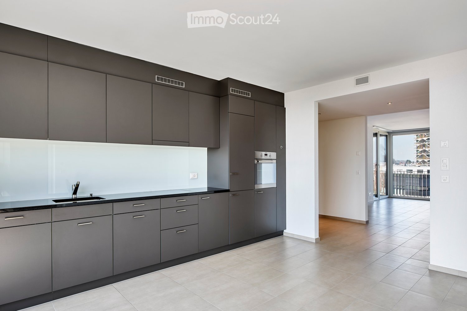 Modern kitchen with grey cabinets, a sink, and an oven, leading to a tiled balcony area with a view.