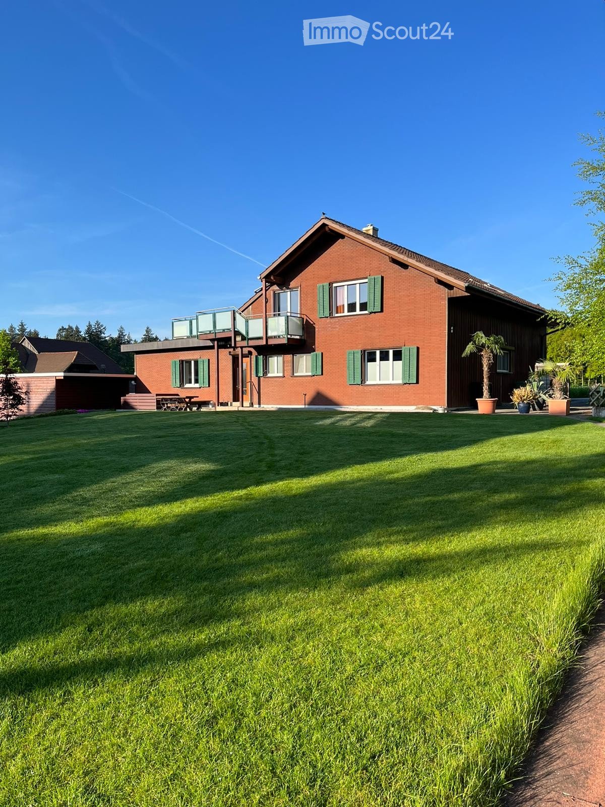 Two-story red brick house with green shutters, large windows, and a balcony on the upper level. The house is surrounded by a well-maintained lawn and features potted plants near the entrance.