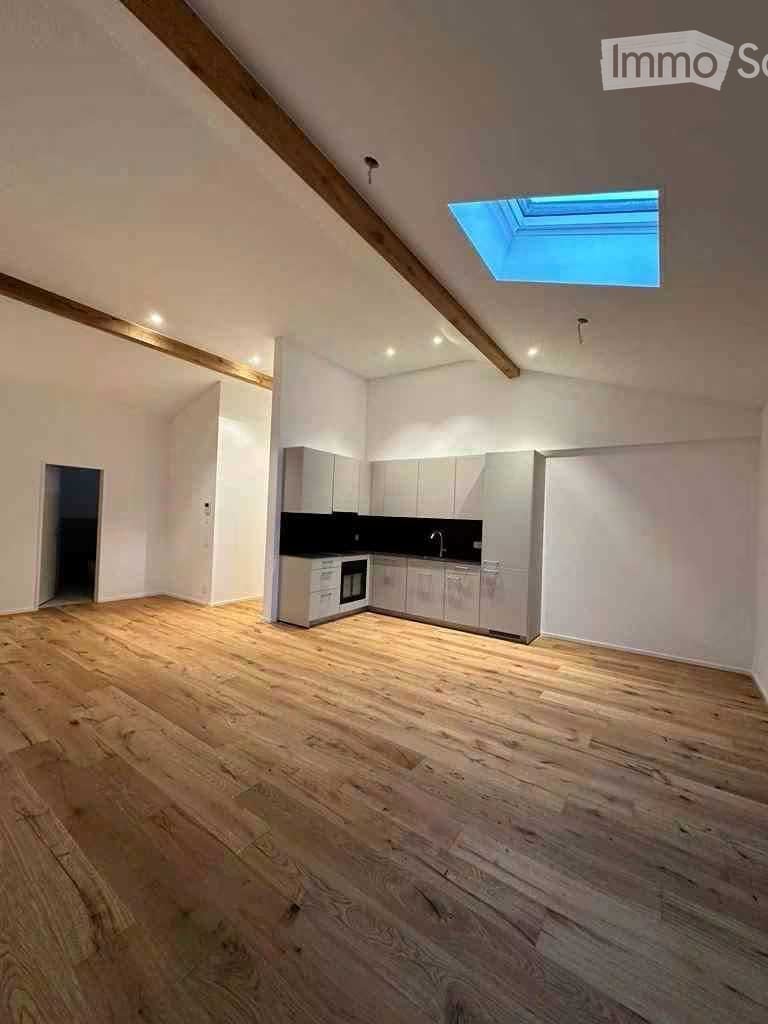 Empty room with wooden flooring, white walls, and a small kitchen area with cabinets and appliances.