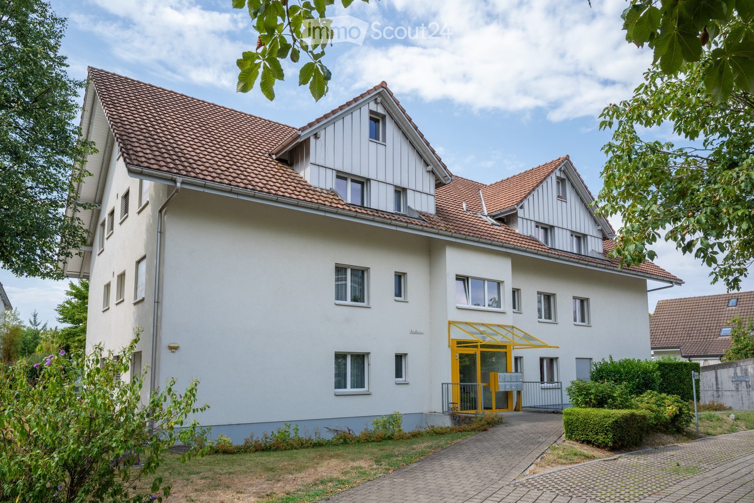 Two-story house with white exterior, red-tiled roof, multiple windows, and a yellow entrance with a small covered porch.